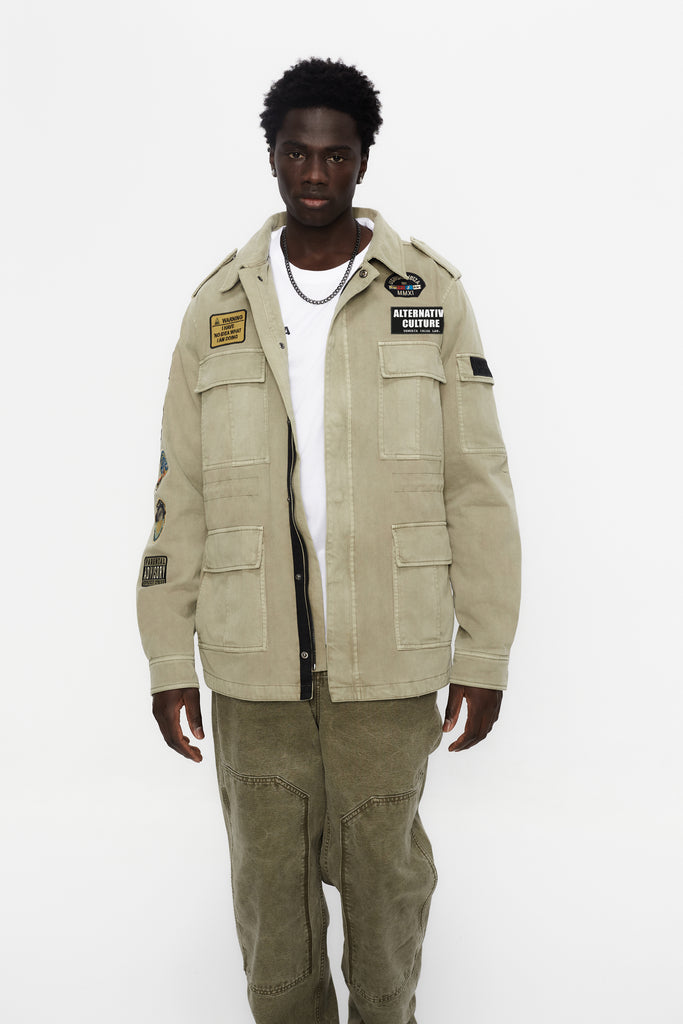 Washed twill military jacket with shirt collar and long sleeves with adjustable cuff with button. Central zip closure with adjustable drawstring at the waist. Patch pockets and flap on the front and sleeve. Combination of patches and embroidered application on front and back. Custom label detail on the back.